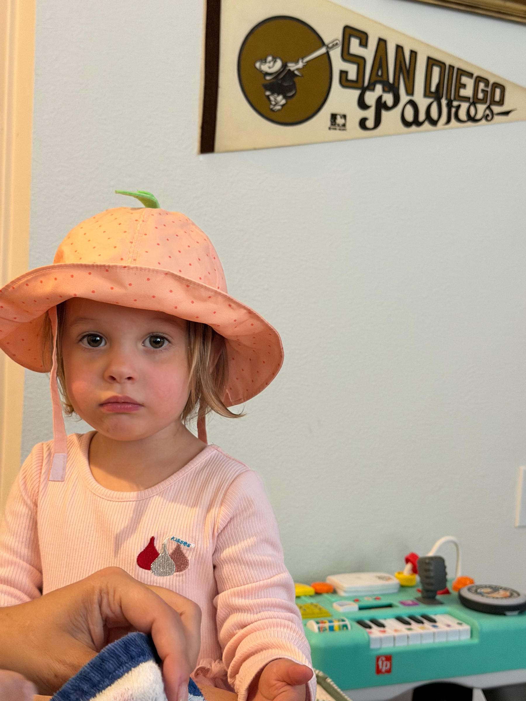A young child wearing a peach-colored hat and light pink shirt stands in a room with a San Diego Padres pennant on the wall and a toy piano in the background.